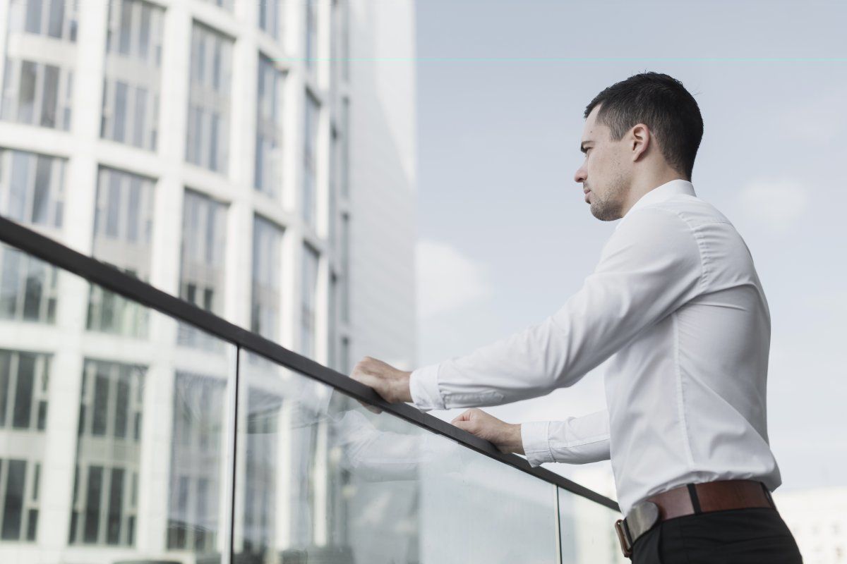 Retrouver sa légitimité professionnelle après un épuisement – Coaching Stressmanager.fr Homme en chemise accoudé à une rambarde, regardant les buildings en arrière-plan, symbole de remise en question professionnelle et de recherche de sens.